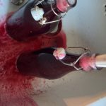 glass bottles full of red liquid in a white sink with red liquid sprayed out of them. a white plastic funnel is out of focus in one of the bottles to the right