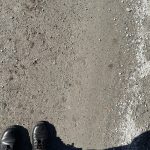 a pair of feet wearing boots with standing at the water's edge at the beach in the sand and there is a shadow to the right